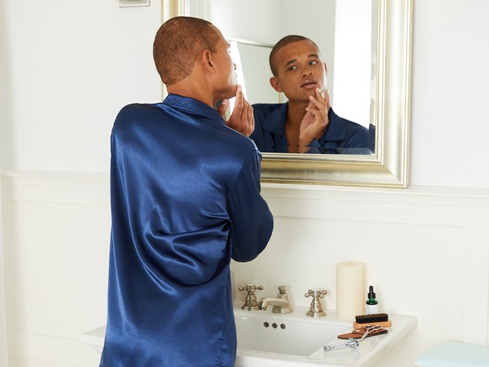 Person in blue silk pajamas looking in the mirror in the bathroom and touching their face